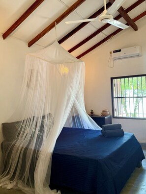 A bedroom with a bed covered by a white canopy, a ceiling fan, an air conditioning unit, a window with bars, and a small table with a plant and a decorative item. - Peaceful, private self contained villa. Just a few steps from the beach! (Tangalle)