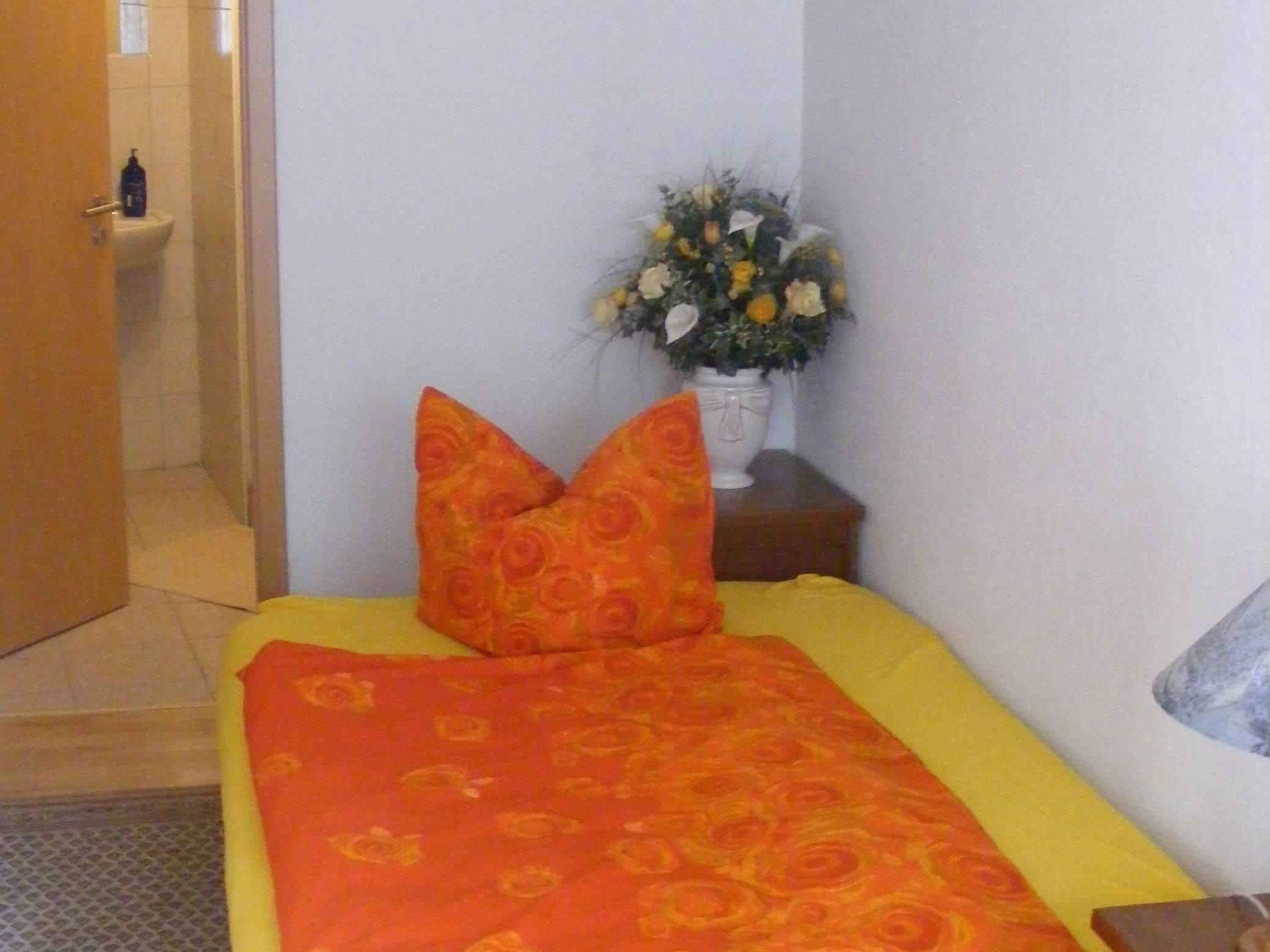 A small, neatly arranged bedroom with a bed, pillows, a nightstand, and a vase of flowers.