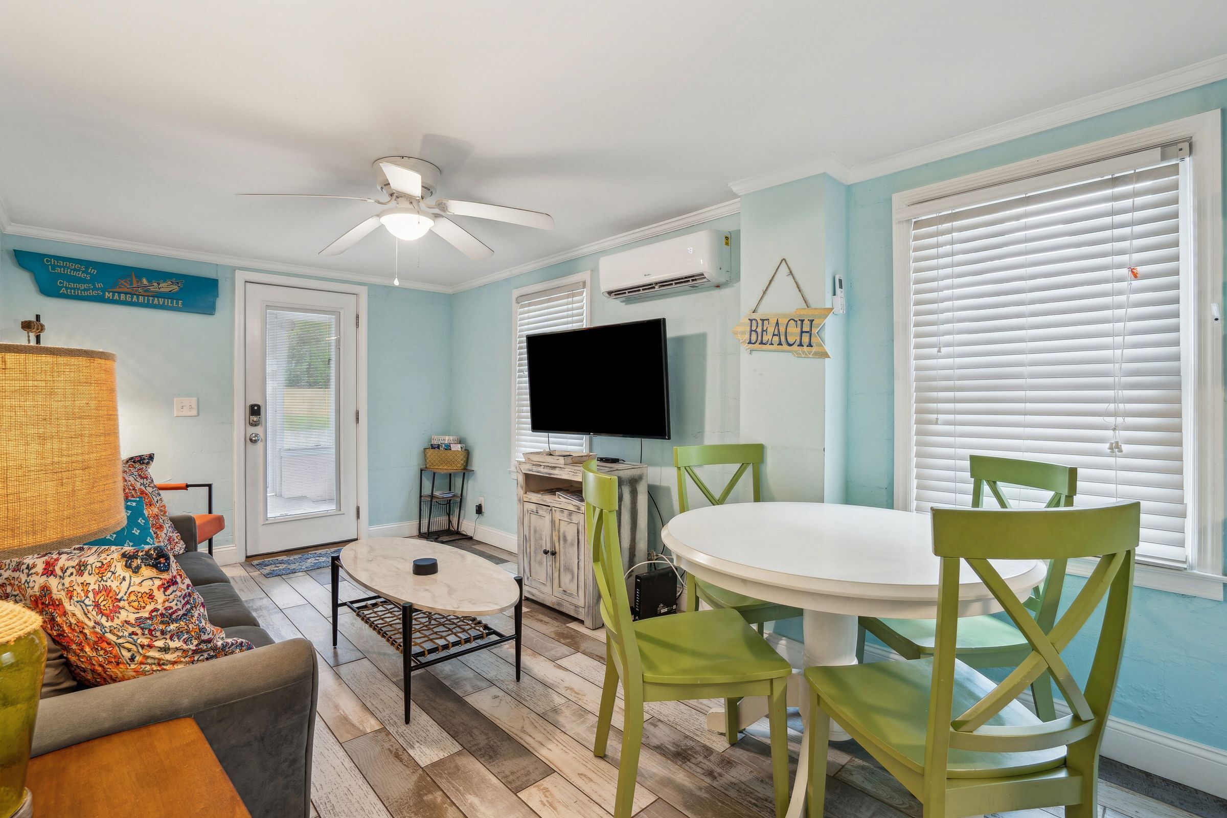 A dining area with a white table and green chairs, a ceiling fan, and a television on the wall.
