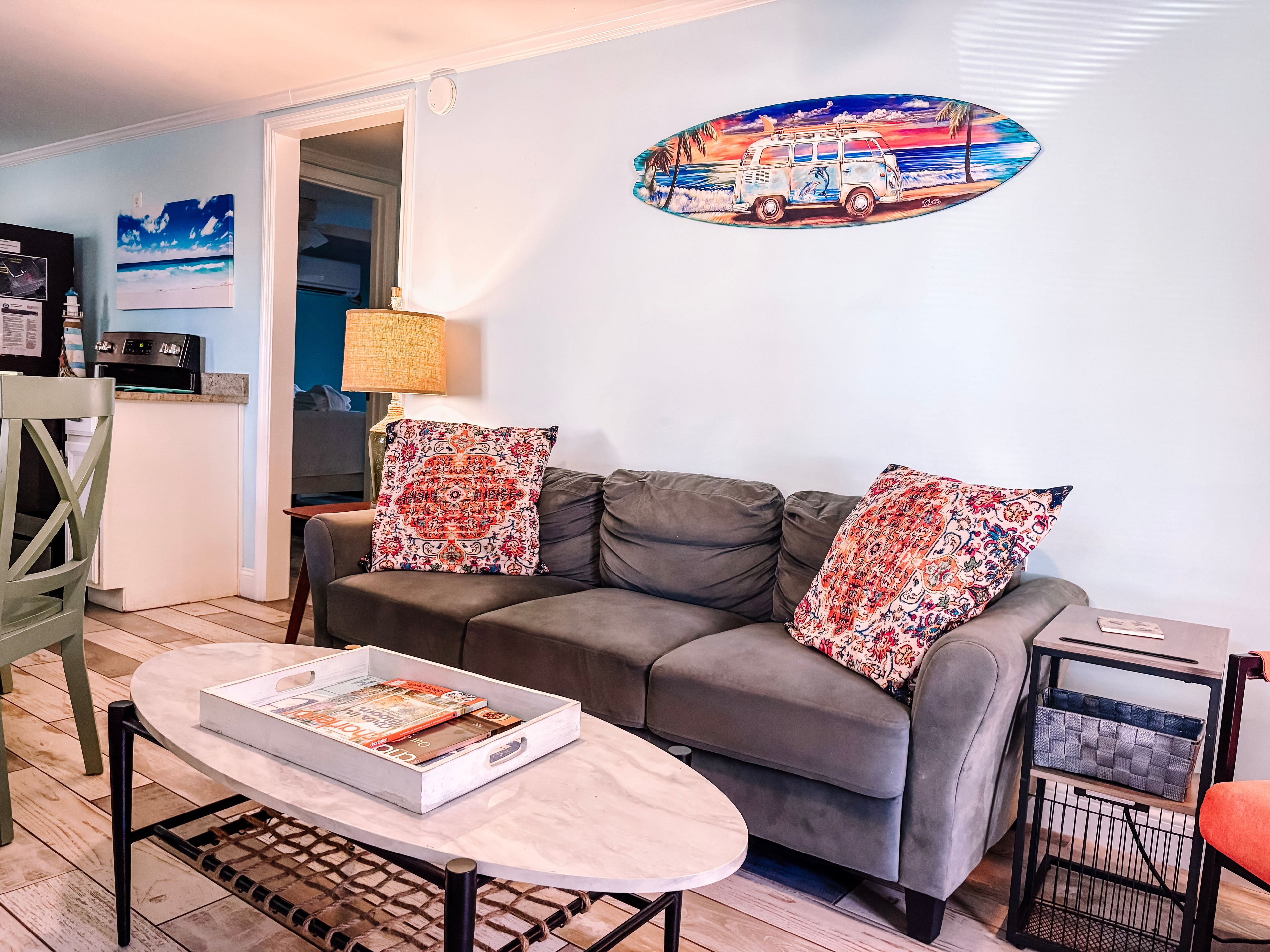 A living room with a grey sofa, two patterned cushions, a coffee table with a magazine, a lamp, and a wall art of a van.