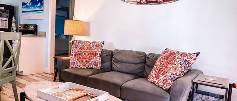 A living room with a grey sofa, two patterned cushions, a coffee table with a magazine, a lamp, and a wall art of a van.