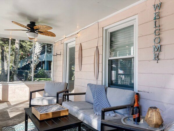 A sunlit porch with a ceiling fan, a sofa with patterned cushions, a chessboard, and a bottle of champagne. - Short Walk to Shops, Restaurants & Beach Access,Pet Friendly, Private fenced in yard (Folly Beach)