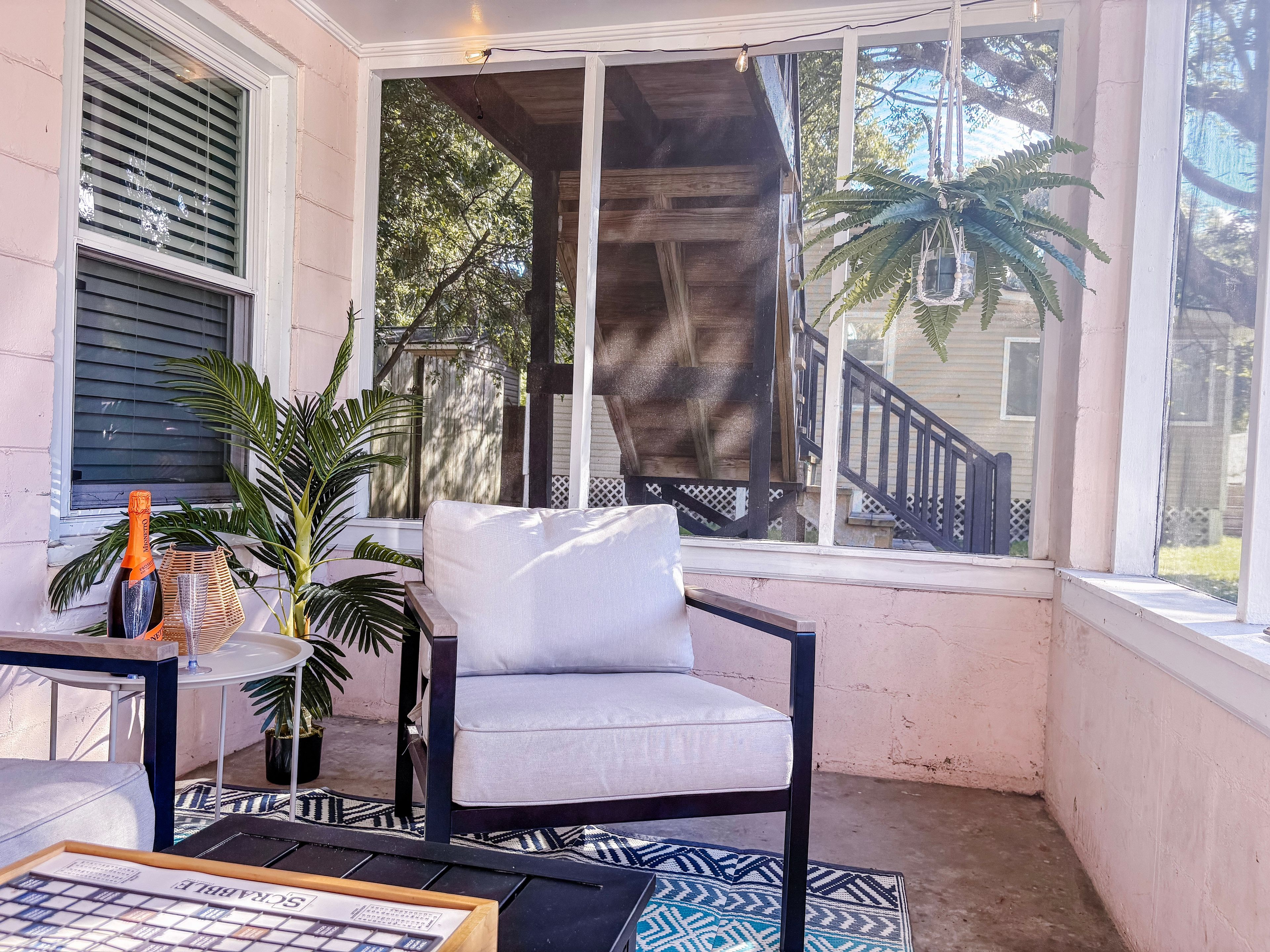 A covered porch with a white cushioned chair, a small table with a magazine, and a bottle of champagne.