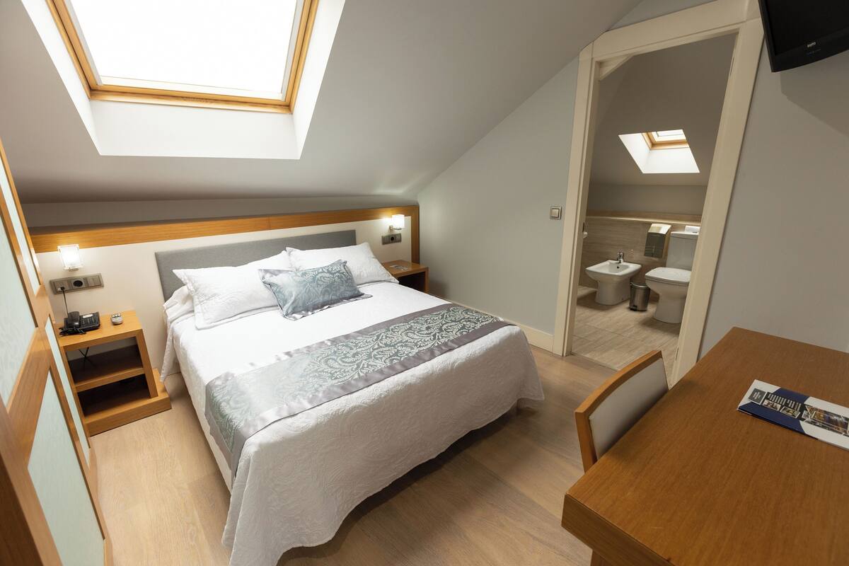 A bedroom with a skylight, a bed with a patterned bedspread, bedside tables with lamps, a wooden nightstand, and a wooden chair.