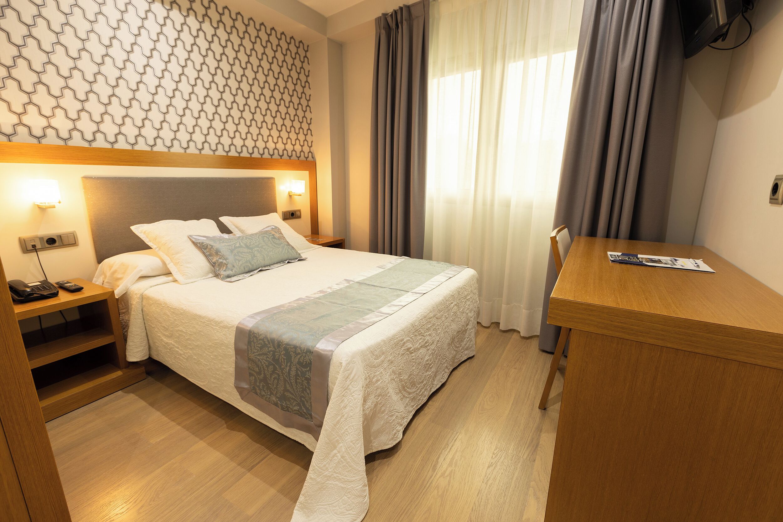 A hotel room with a neatly made bed, a wooden desk, and a patterned wall.