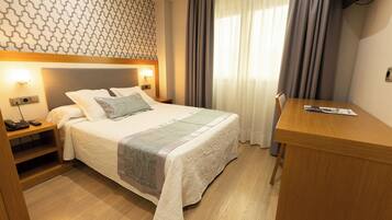 A hotel room with a neatly made bed, a wooden desk, and a patterned wall.