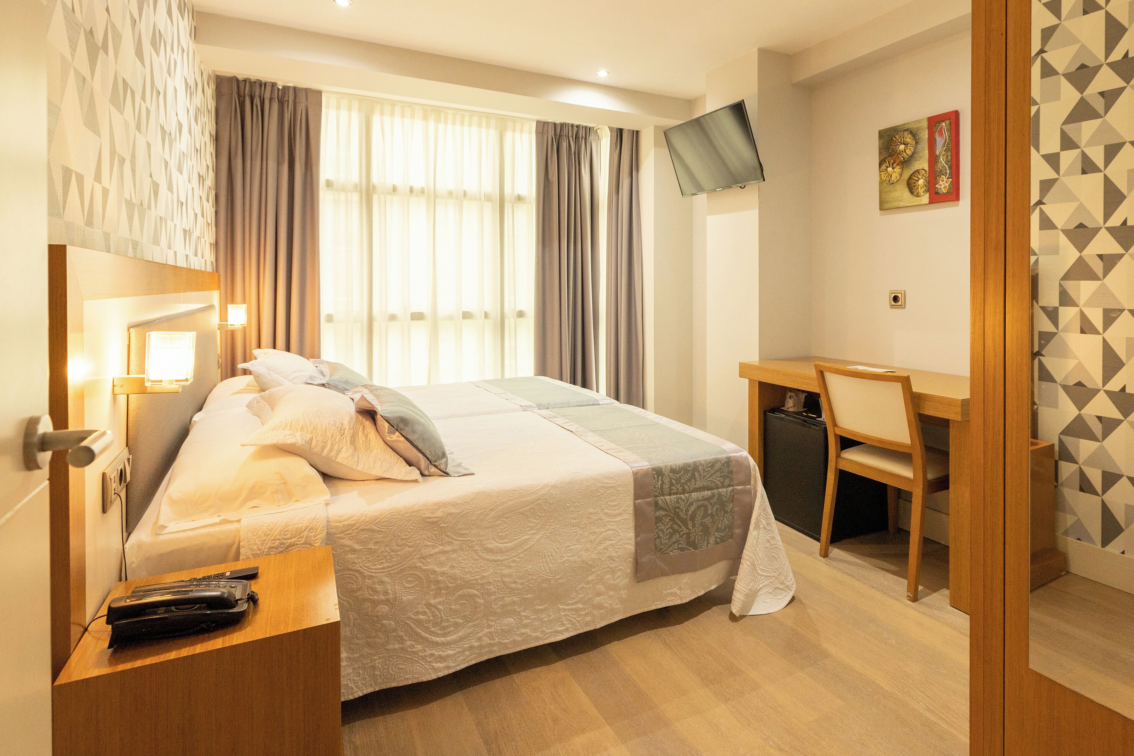 A hotel room with a bed, bedside table, desk, chair, TV, and curtains.