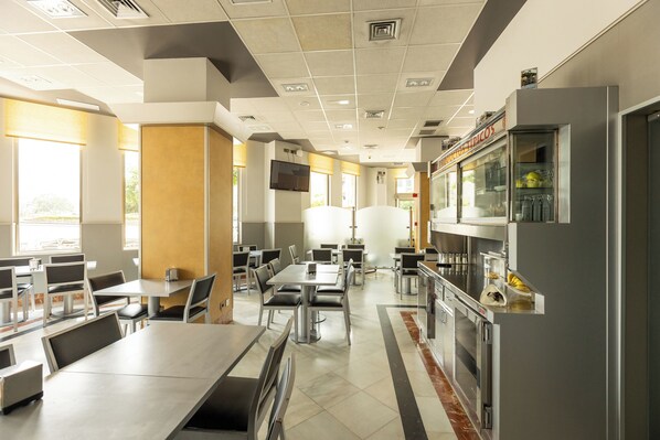 A modern cafeteria with stainless steel tables and chairs, a refrigerator with beverages, and a television mounted on the wall.