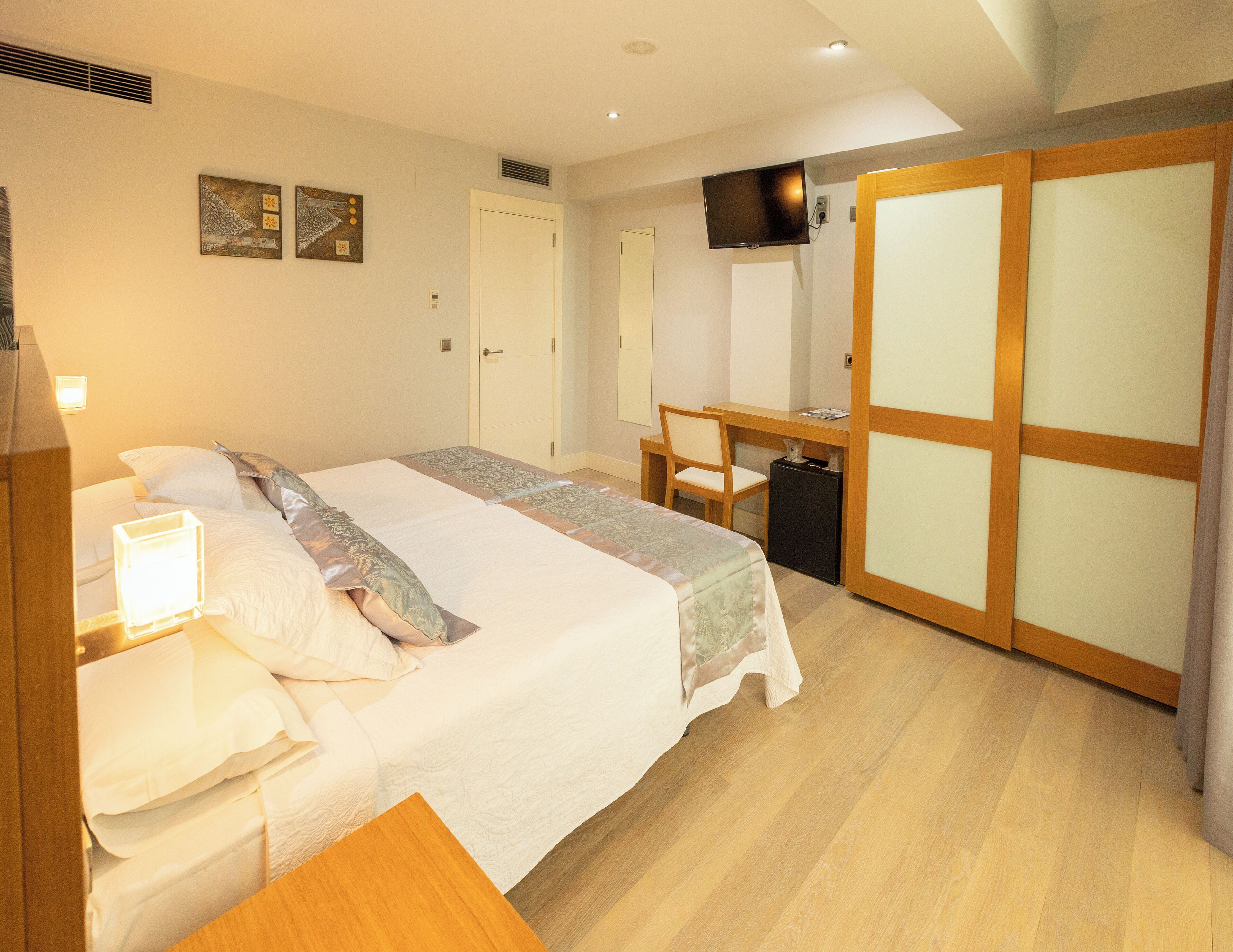 A hotel room with a bed, desk, chair, TV, wardrobe, and artwork on the wall.