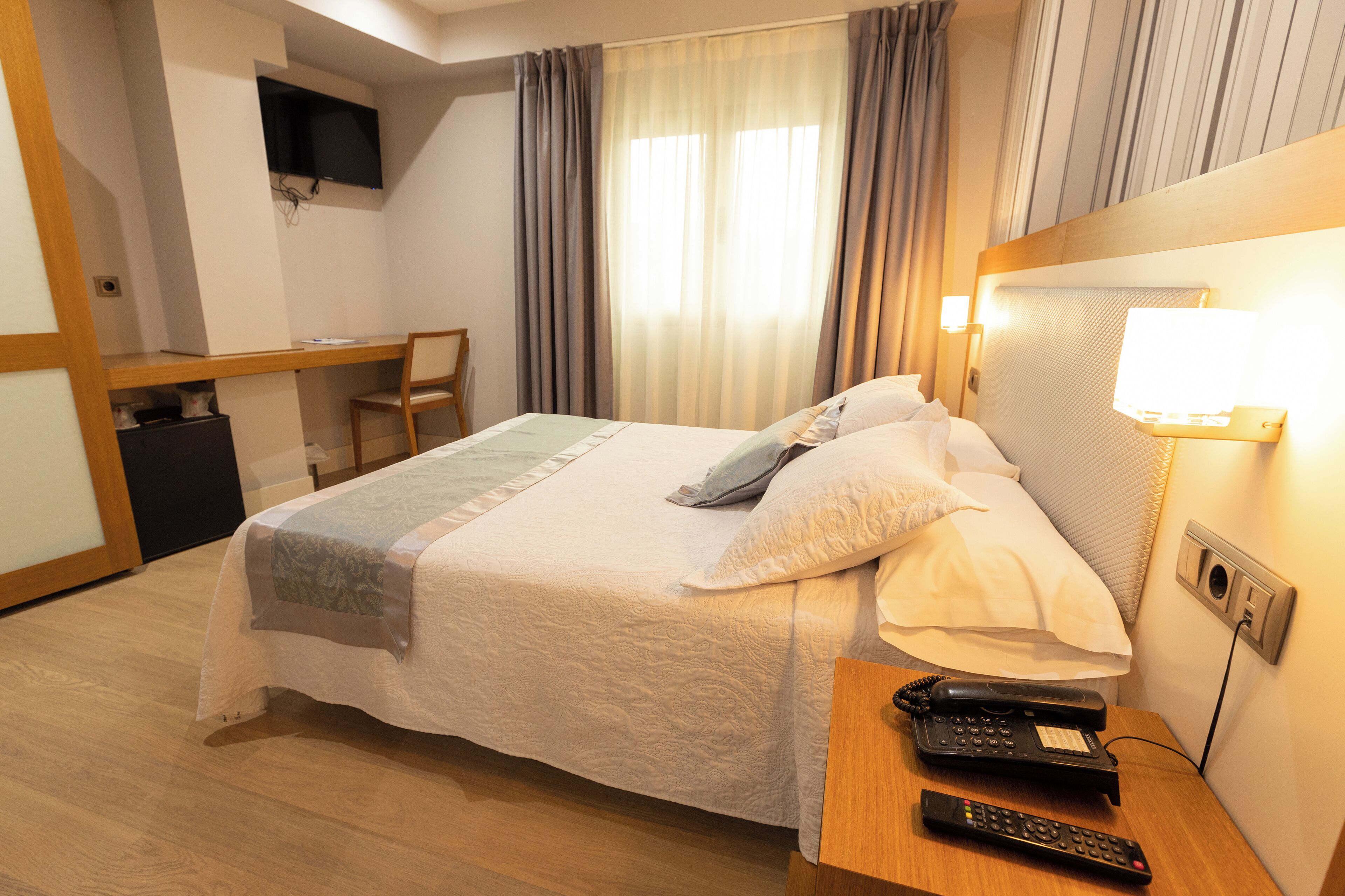 A hotel room with a bed, a desk, a chair, a TV mounted on the wall, and a bedside table with a phone and remote control.