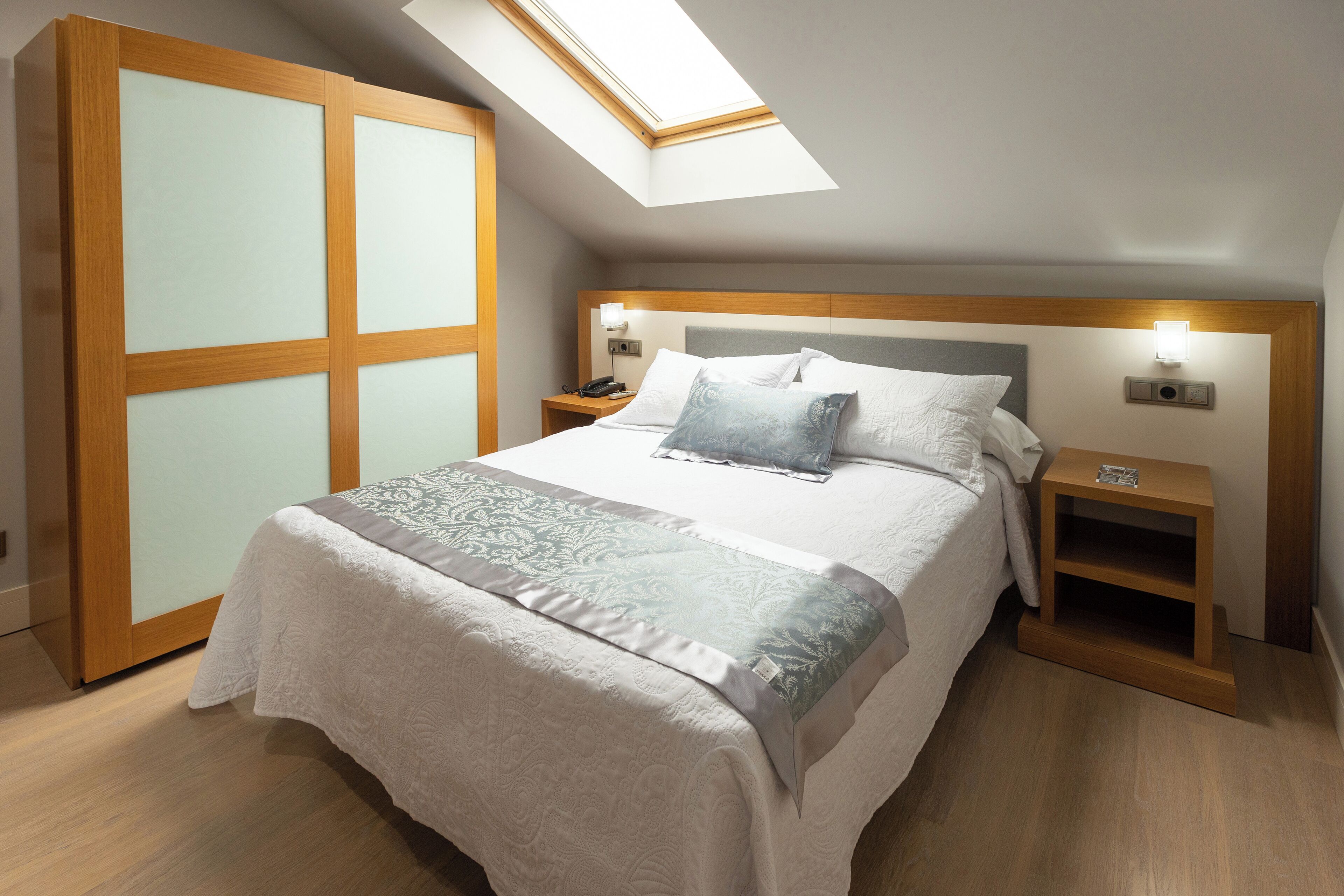 A bedroom with a skylight, a bed with a patterned bedspread, bedside tables with lamps, and a wooden wardrobe.