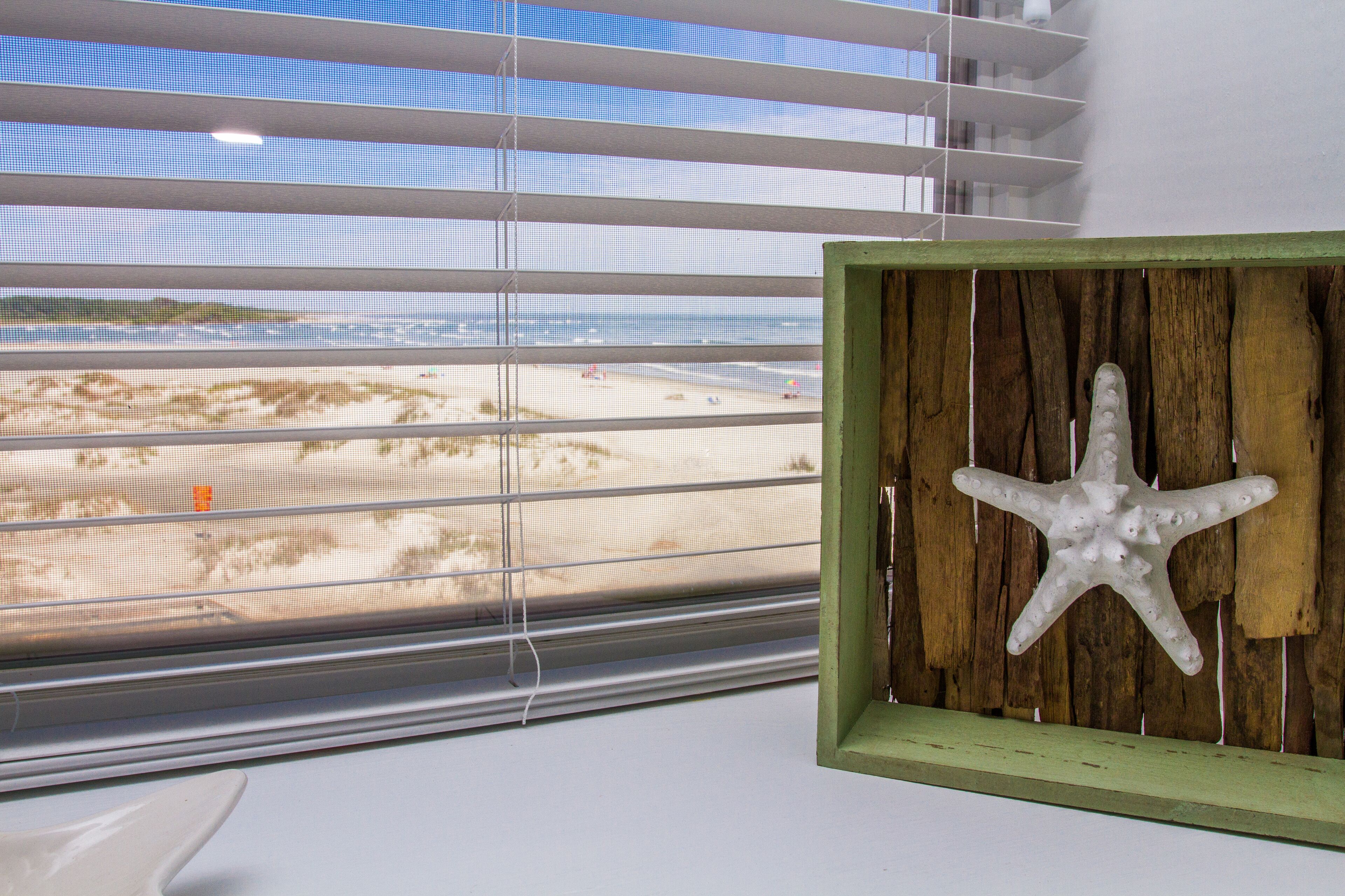 Una ventana con persianas horizontales, un marco de madera con una decoración de estrella de mar, y vista a una playa.
