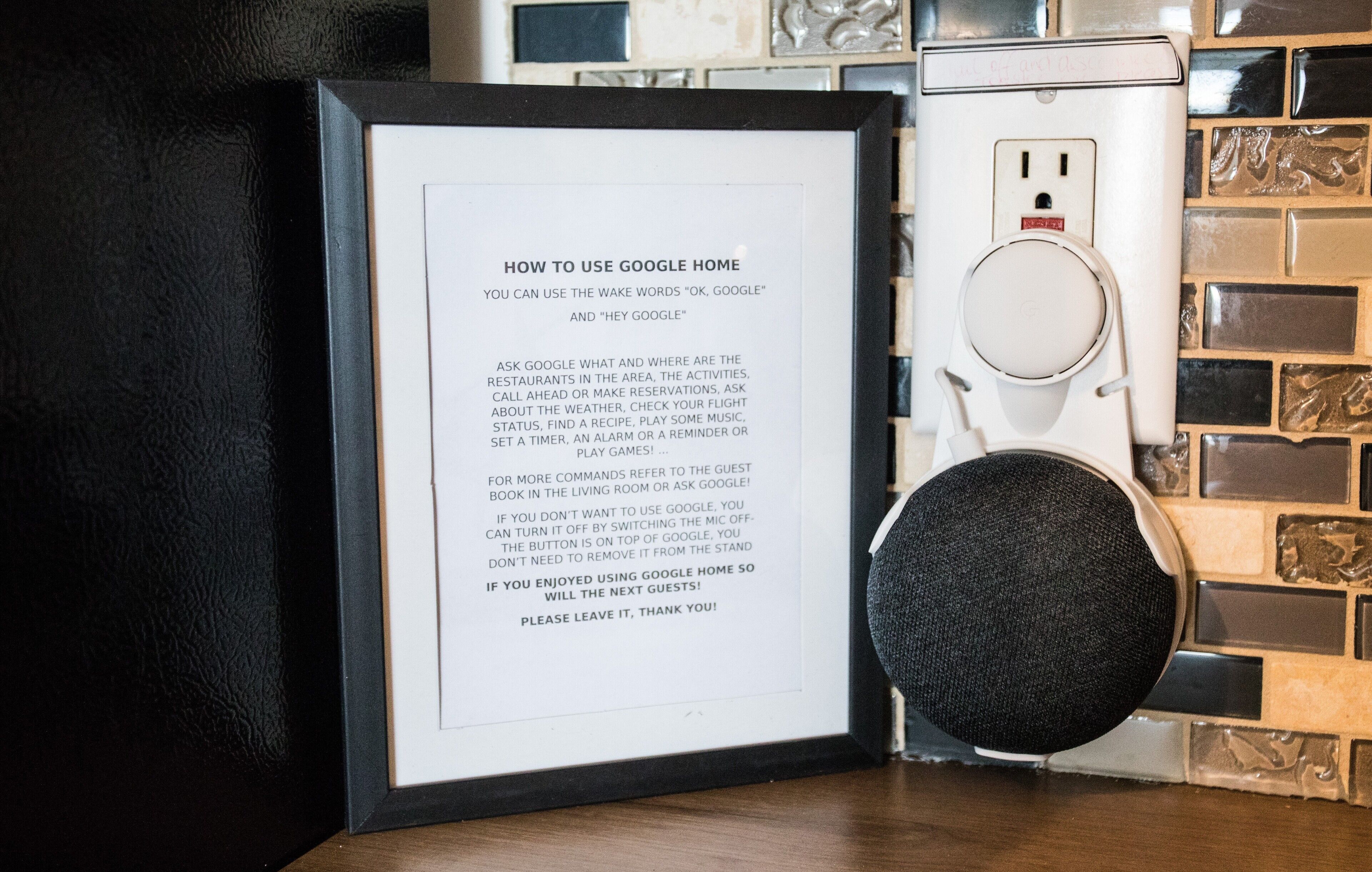Et indrammet instruktionstabel for brug af Google Home, placeret ved siden af en smart højttaler på en bordplade med flisebeklædning.