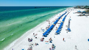Ein Strand mit einer langen Reihe blauer Sonnenschirme und Liegestühlen, einer Mole, die sich ins Meer erstreckt, und Menschen, die die Sonne genießen.