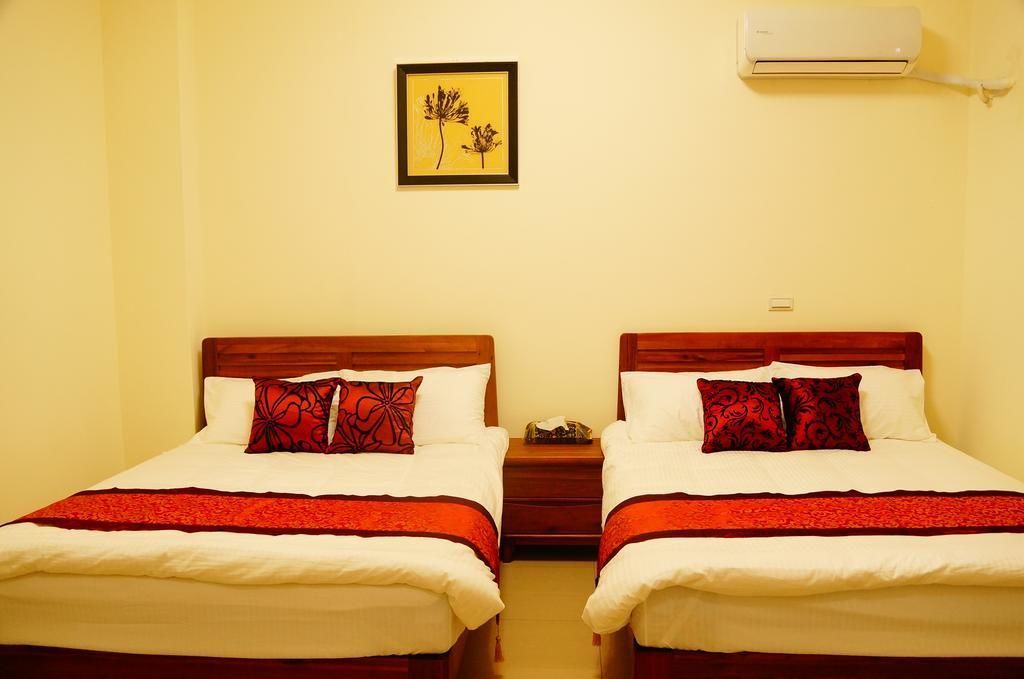 A hotel room with two beds, each with a red and white bedspread and matching pillows. There is a framed picture on the wall above the beds and an air conditioning unit mounted on the wall.