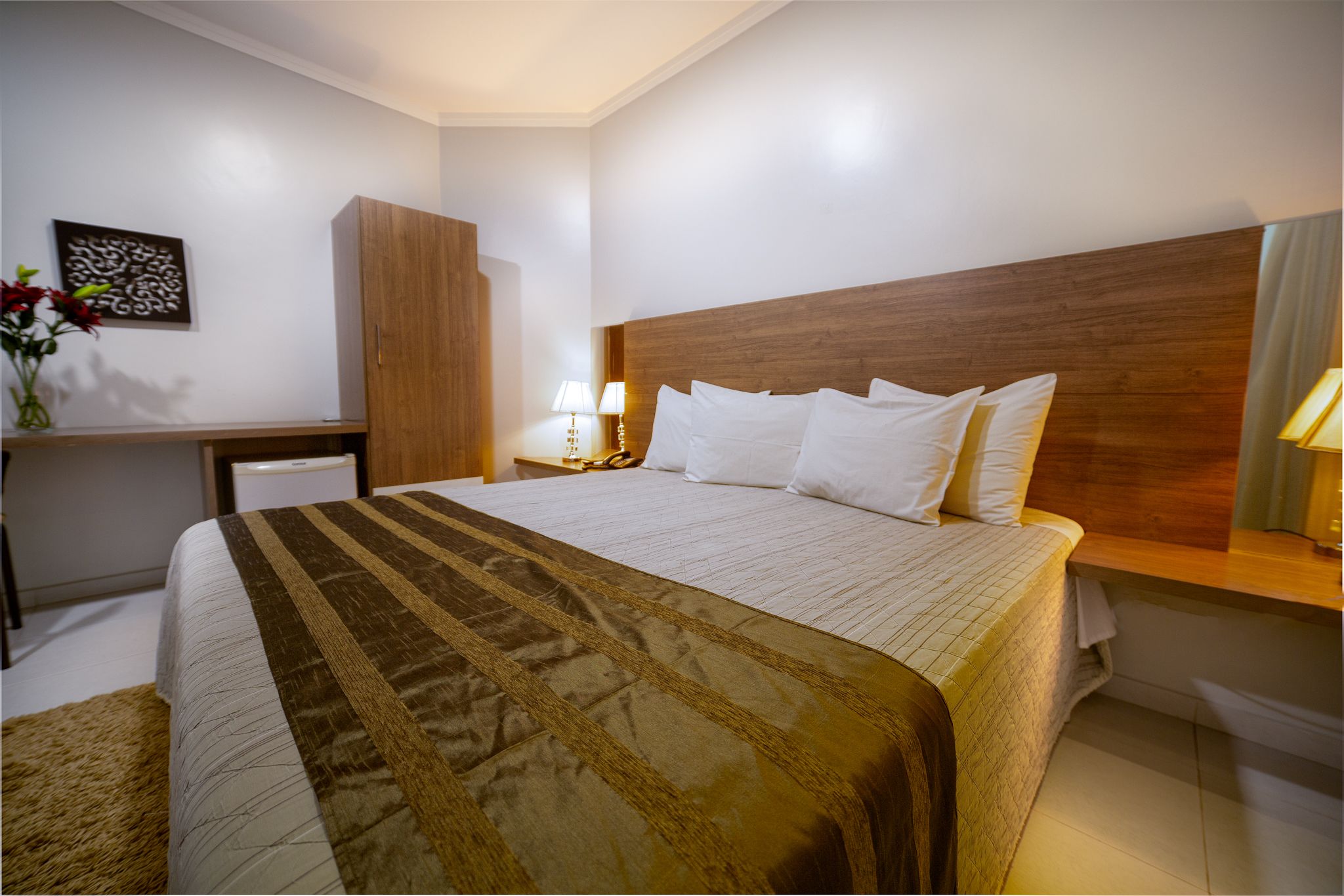A hotel room with a large bed, wooden headboard, bedside table with lamp, and a desk with a chair.