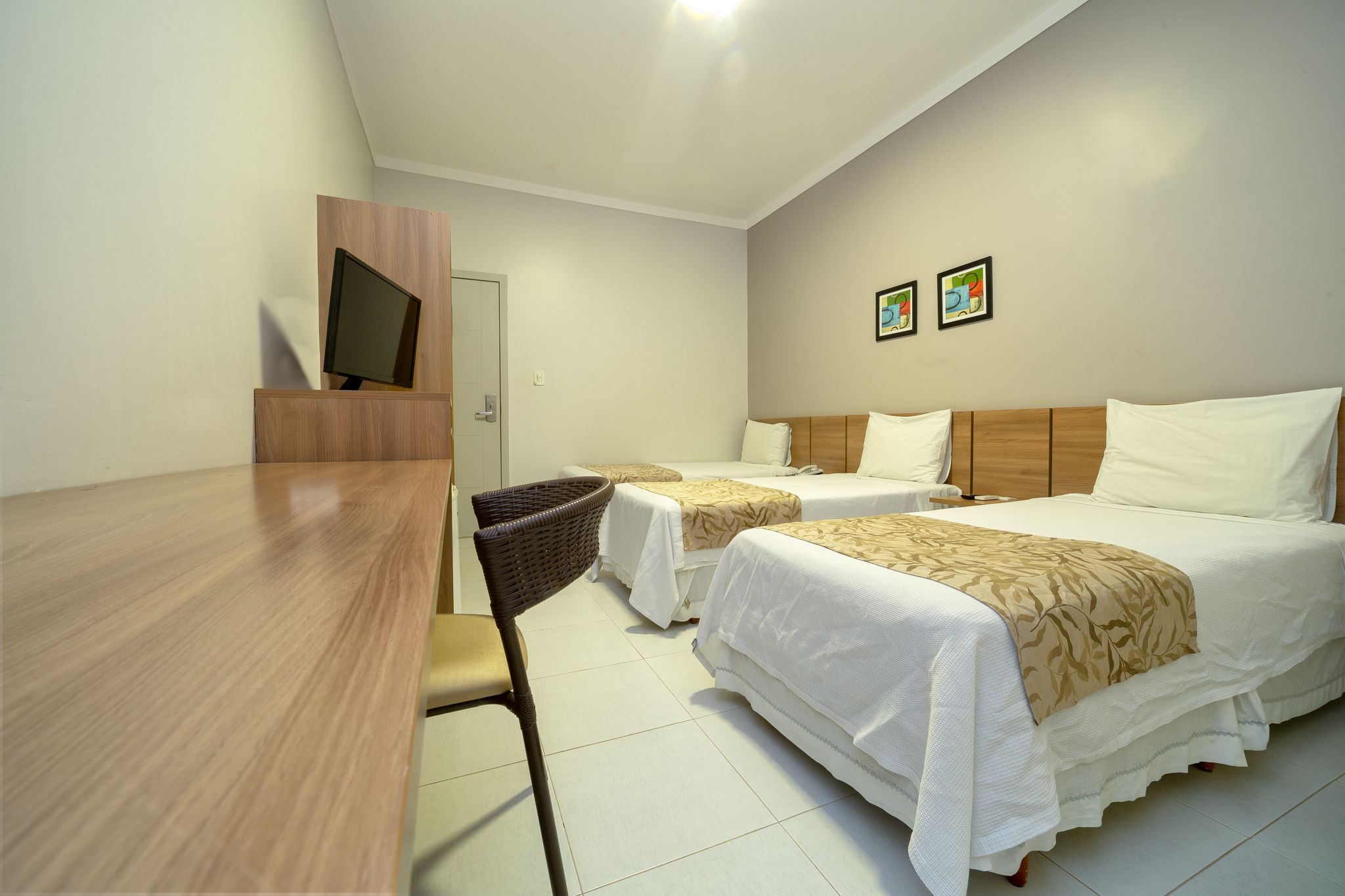 A hotel room with two beds, a wooden desk, a television, and two framed pictures on the wall.