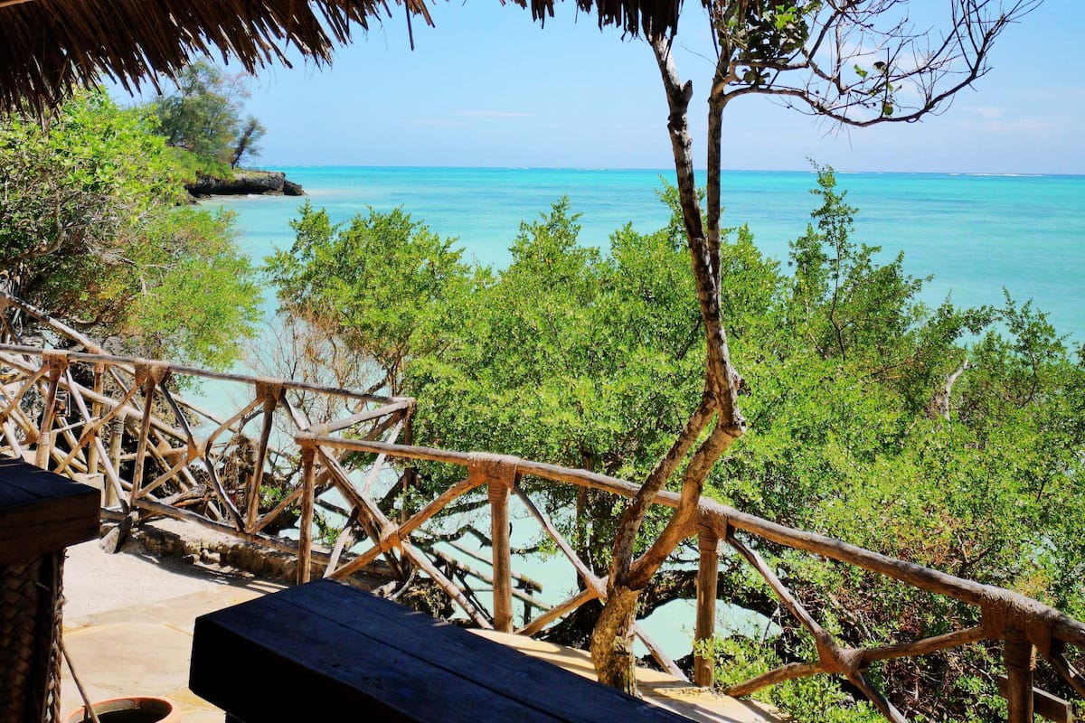 Een houten terras met rieten dak, met uitzicht op turquoise water en weelderige begroeiing.