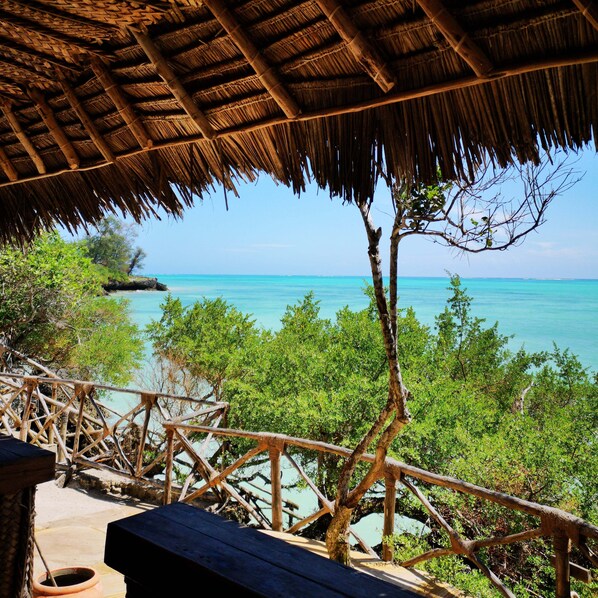 Une terrasse en bois surmontée d’un toit de chaume, offrant une vue sur des eaux turquoise et une végétation luxuriante.