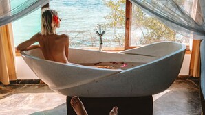 Een slaapkamer met een groot raam met uitzicht op de zee, een badkuip en een bed met een persoon die een smartphone gebruikt.