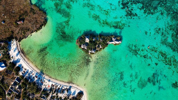 Luchtbeeld van een tropisch eiland met helder turquoise water, een kleine groep gebouwen omgeven door weelderige begroeiing, en een zandstrand met parasols.