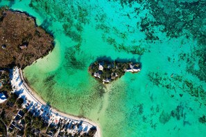 Une vue aérienne d’une île tropicale aux eaux turquoise et limpides, avec un petit groupe de bâtiments entourés d’une végétation luxuriante et une plage de sable bordée de parasols.