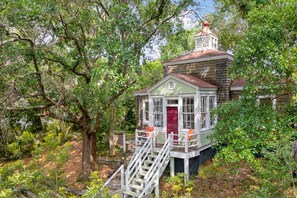 A small, two-story house with a red door and white trim, surrounded by lush greenery.