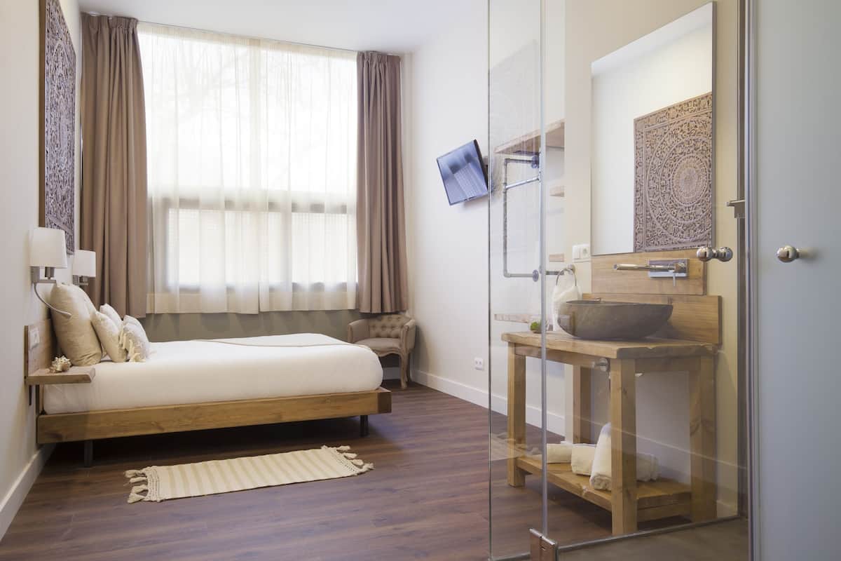 Habitación de hotel moderna con una cama de madera, cabina de ducha de vidrio, tocador de madera con un lavabo grande y un televisor de pantalla plana montado en la pared.