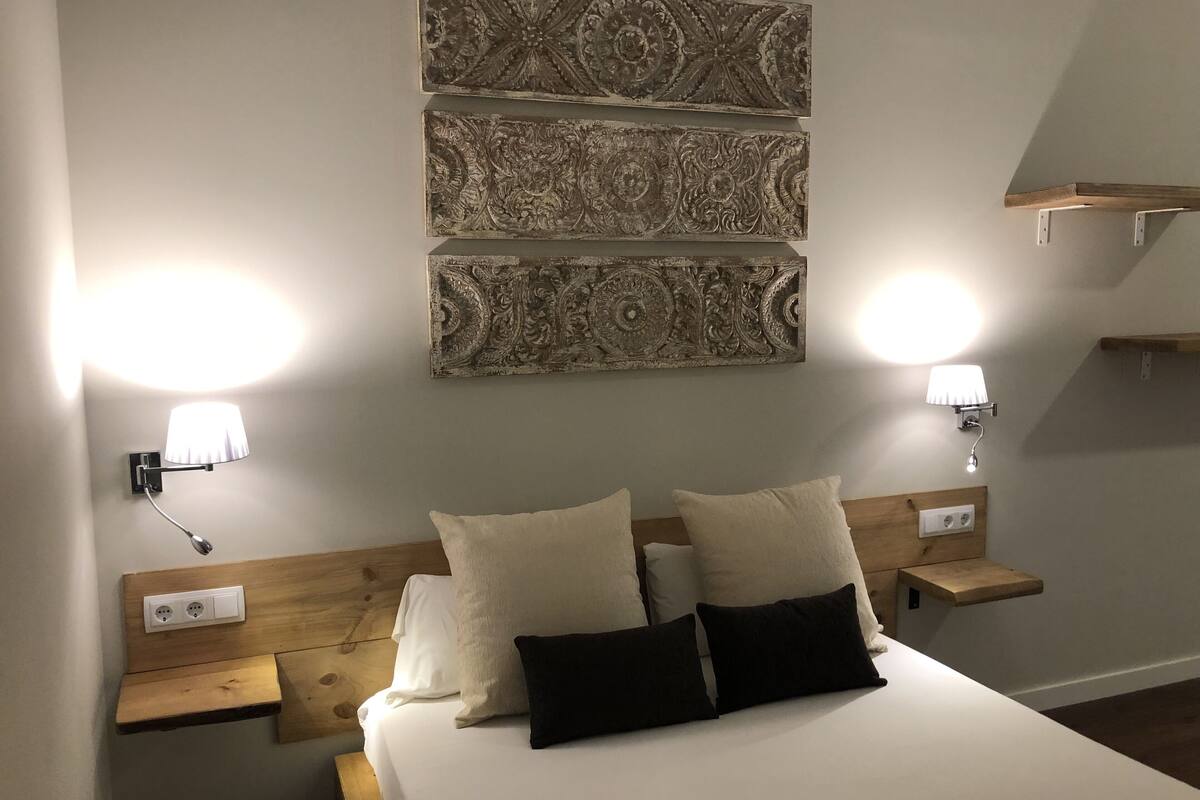 Un dormitorio con cabecera de madera, una cama con almohadas blancas y negras, y dos mesitas de noche fijadas a la pared con lámparas.