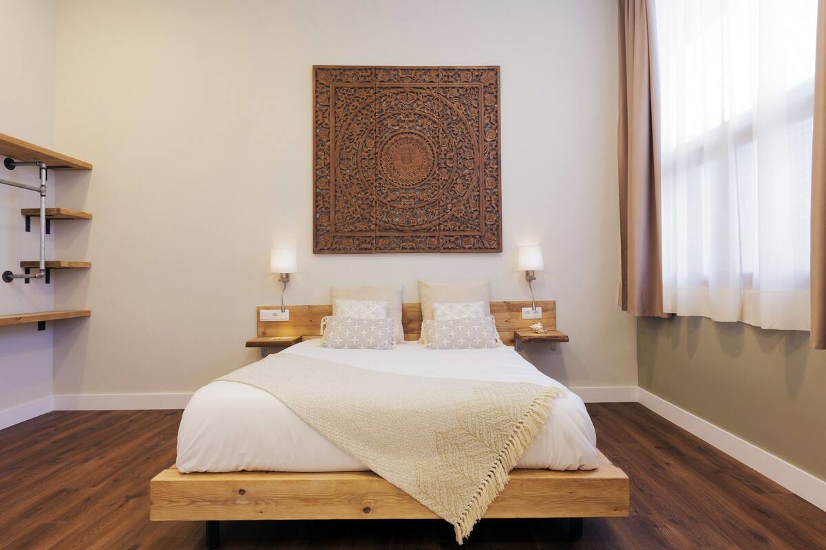 Un dormitorio con cama de madera, ropa de cama blanca, un adorno en la pared, lámparas de noche y una ventana con cortinas.