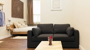 Un salon moderne avec un canapé gris foncé, une table basse en bois et un lit recouvert d’une couette blanche.