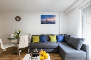 A modern living room with a grey sectional sofa, a dining table with chairs, and a cityscape painting on the wall.