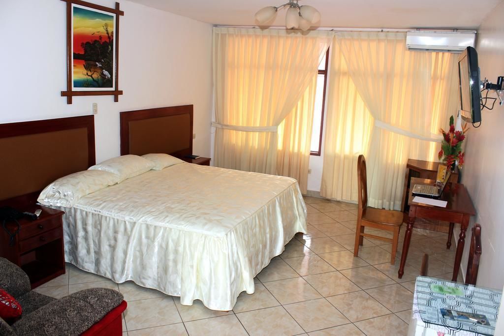 A hotel room with a large bed, a desk with a chair, a small table, and a window with curtains.