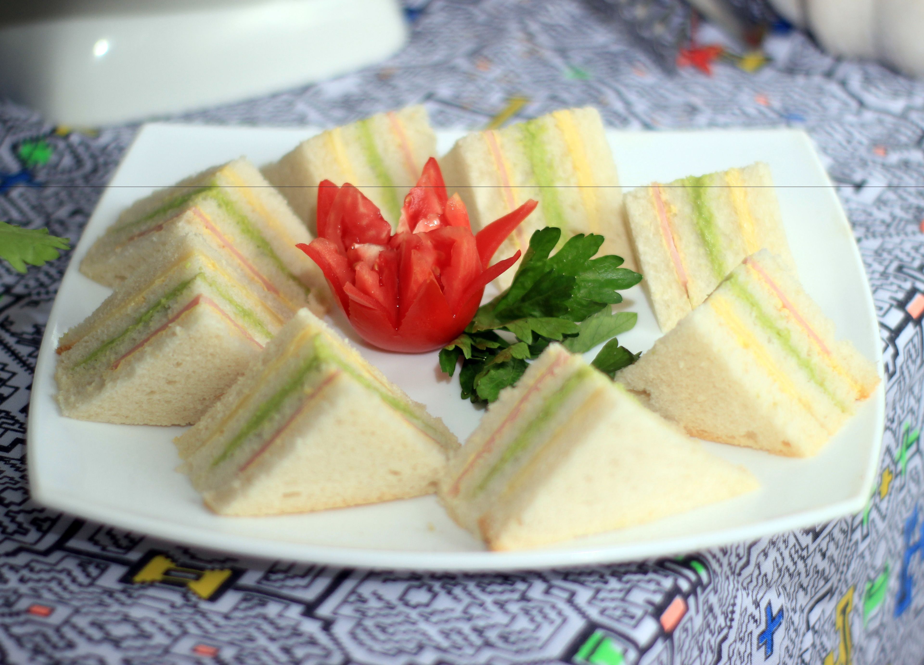 A plate of triangular sandwiches arranged in a fan shape, garnished with a tomato flower and parsley, on a patterned tablecloth.