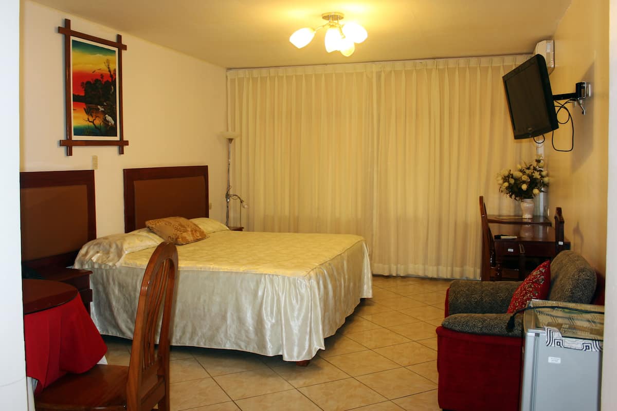 Une chambre d’hôtel avec un lit, une chaise, une table, un téléviseur et une fenêtre avec des rideaux.
