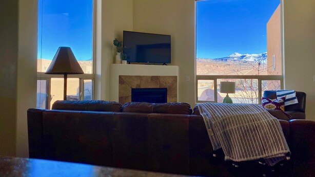 A living room with a fireplace, a television mounted above it, and large windows offering a view of a mountainous landscape. - 10A1 Close to Pool, Views, Huge Patio, Sleeps 8 (Moab)