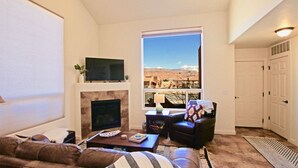 A living room with a fireplace, a television, a sofa, and a view of a mountainous landscape through a large window. - 10A1 Close to Pool, Views, Huge Patio, Sleeps 8 (Moab)