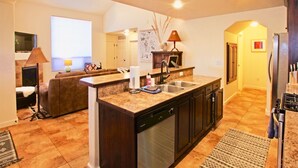 A modern kitchen with a built-in dishwasher, a sink, and a countertop. There is a dining area with a couch and a lamp. The kitchen has a window and a door leading to another room. - 10A1 Close to Pool, Views, Huge Patio, Sleeps 8 (Moab)