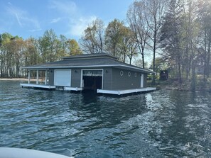 A houseboat with a covered porch and a garage, floating on a body of water surrounded by trees.