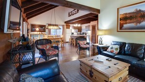 A spacious living area with a wooden coffee table, leather sofas, and wicker chairs. A kitchen is visible in the background with wooden cabinets and a hanging light fixture. - Basecamp for endless adventure, perfect Black Butte location - Hot Tub! (Sisters)
