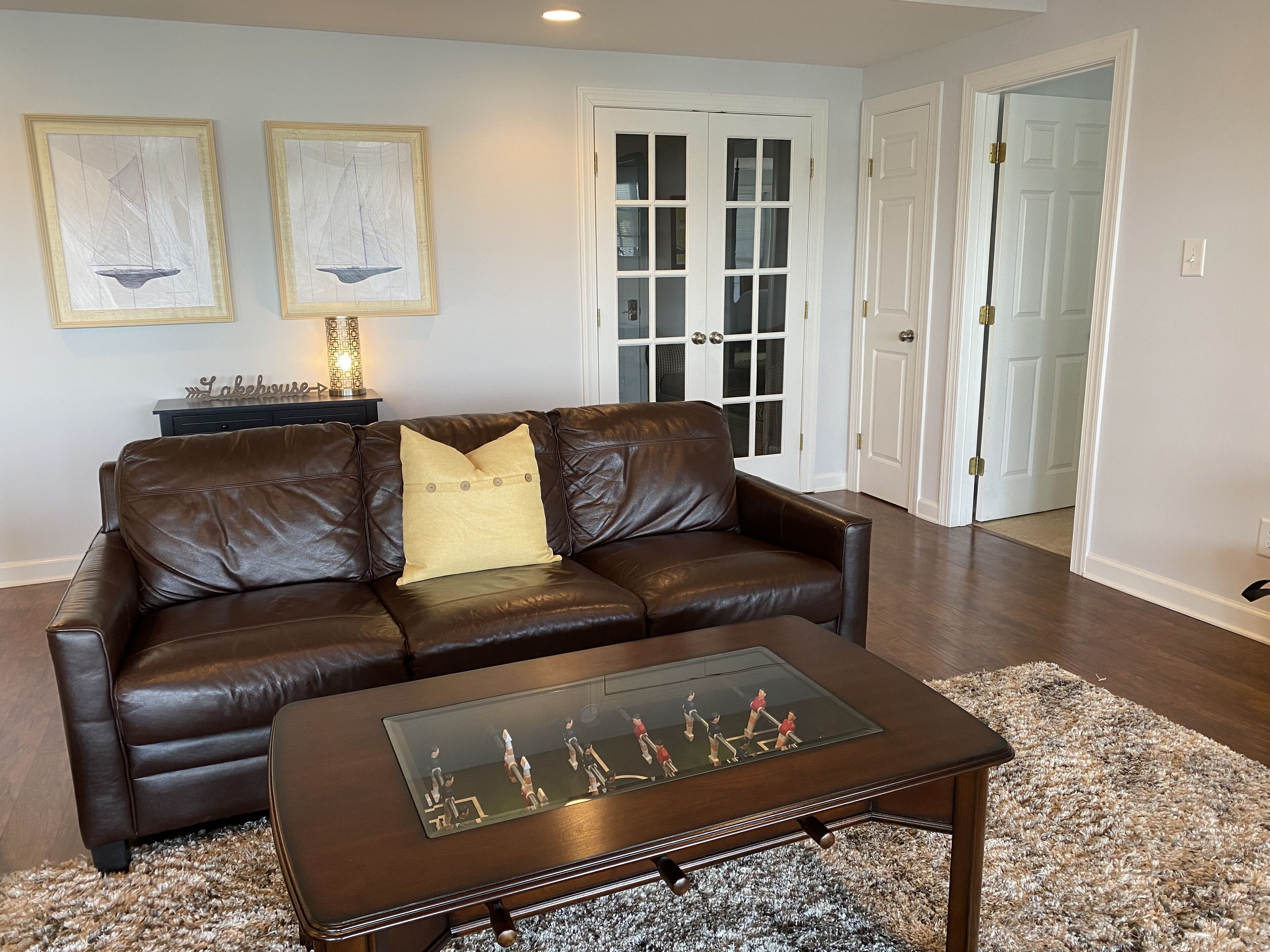A living room with a brown leather sofa, a glass-top coffee table, and two framed sailboat artworks on the wall.