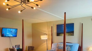 En seng med himmelseng med mørkeblåt sengetæppe, trægulv, et skrivebord med stol, et fjernsyn og et natbord med lampe.