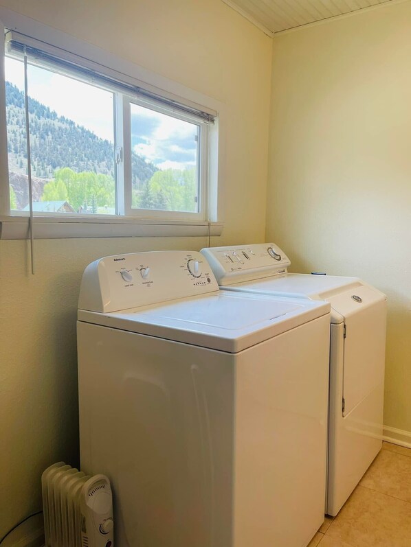 A laundry room with a front-loading washer and dryer, a window with a view of a forested area, and a radiator. - Gorgeous Studio home, Fenced yard, Open Yr Rnd, Dog Friendly. (Lake City)