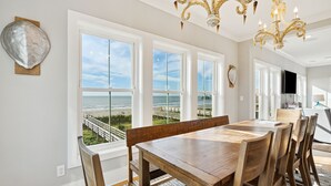 Un espace repas avec une grande table et des chaises en bois, de grandes fenêtres donnant sur la plage et un lustre.