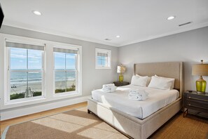 A bedroom with a large bed, two bedside tables, a nightstand, and a view of the ocean.