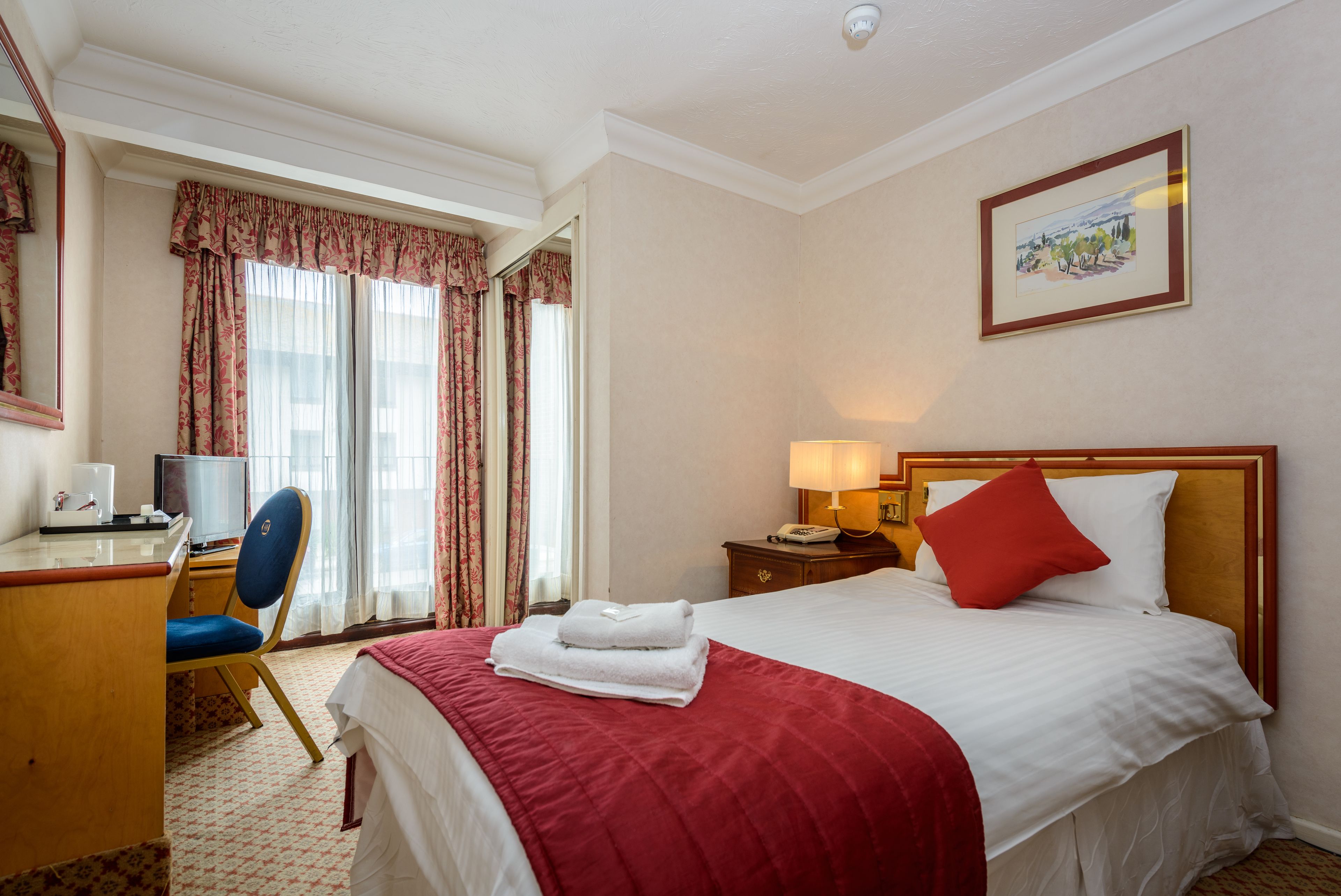 A hotel room with a bed, a desk with a chair, a lamp, and a painting on the wall.