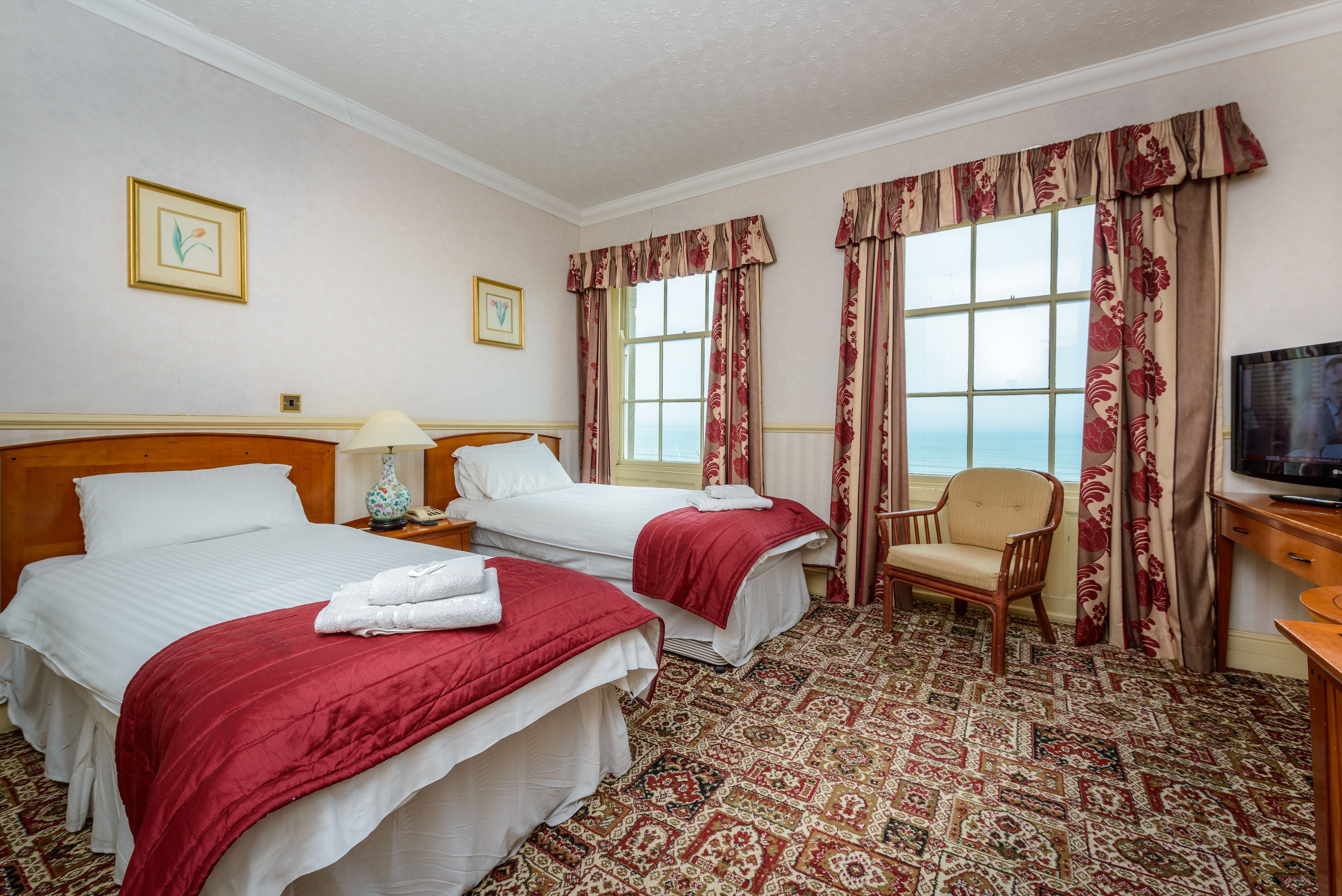 A hotel room with two beds, a chair, a television, and a large window with curtains.