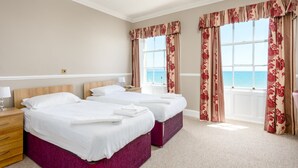 A hotel room with two beds, a wooden nightstand, a lamp, and a view of the sea through large windows with floral curtains. - Russell Hotel (Weymouth)