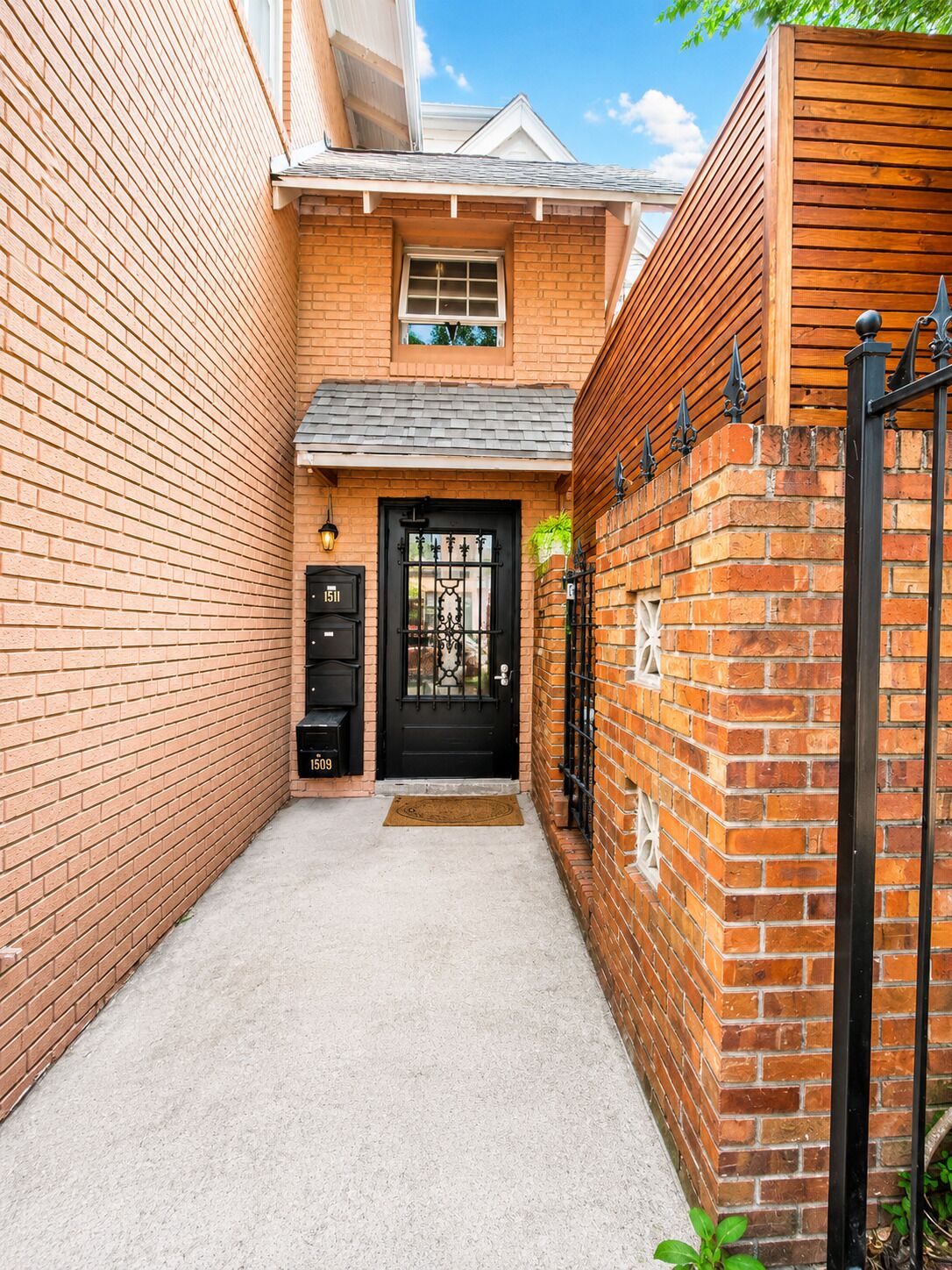 A narrow brick alleyway leading to a black metal gate with a door, flanked by brick walls and a small window above the door.