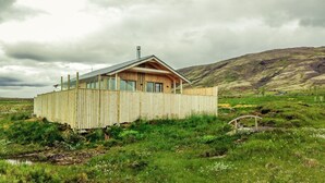 A wooden cabin with a sloped roof, surrounded by a wooden fence, set in a grassy field with a mountainous backdrop.